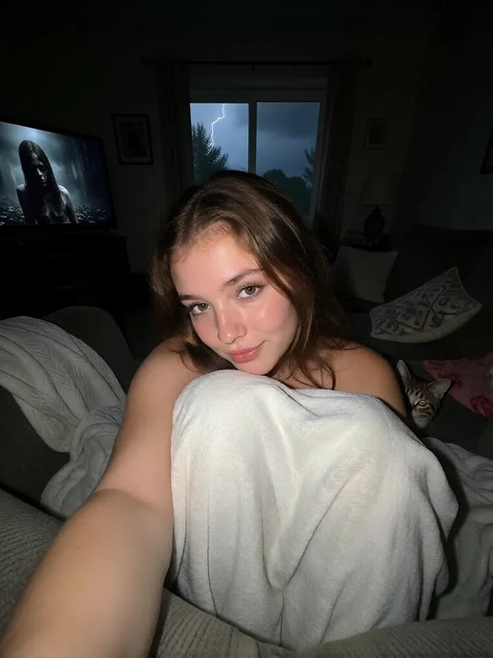 A companion sitting under a blanket on a sofa, with a cat beside her and a storm outside the window